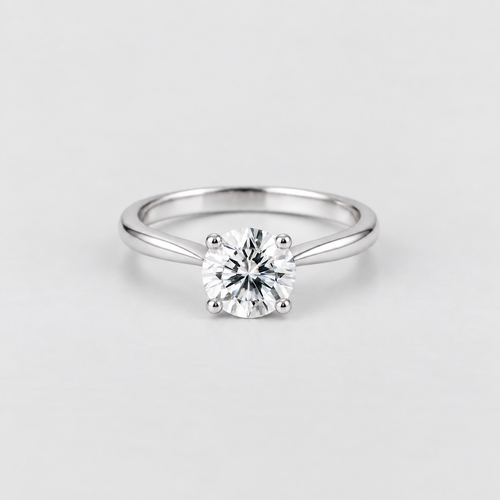 A classic solitaire engagement ring featuring a round brilliant-cut diamond set in a polished four-prong white gold or platinum band, photographed on a clean, light gray background.