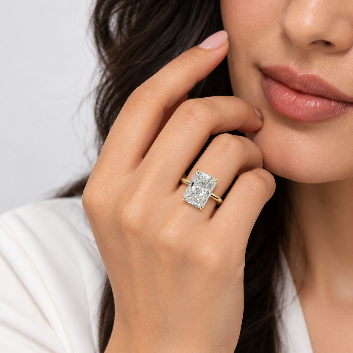 A close-up lifestyle shot of a woman wearing a yellow gold ring with a rectangular emerald-cut diamond. The ring is shown on her finger, with her hand resting near her face against a clean, light gray background.