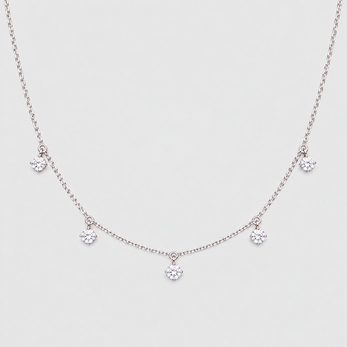 A delicate silver-tone necklace featuring a fine chain with five small, clear crystal flower-like charms spaced along the front, displayed against a light gray background.