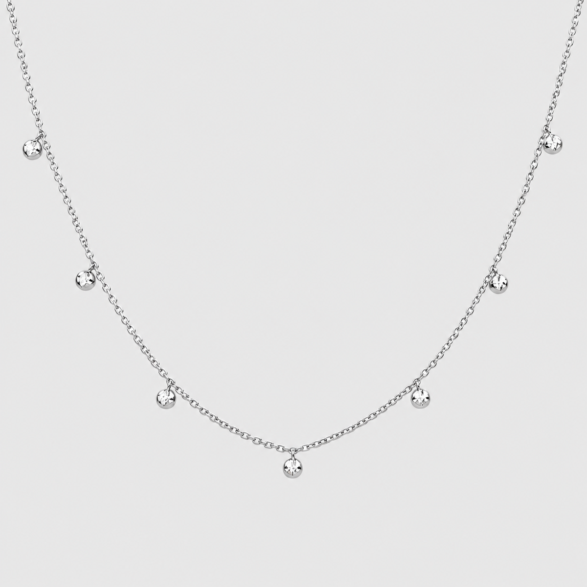 A delicate silver chain necklace featuring seven small, clear round gemstones spaced along the chain, displayed against a solid light gray background.