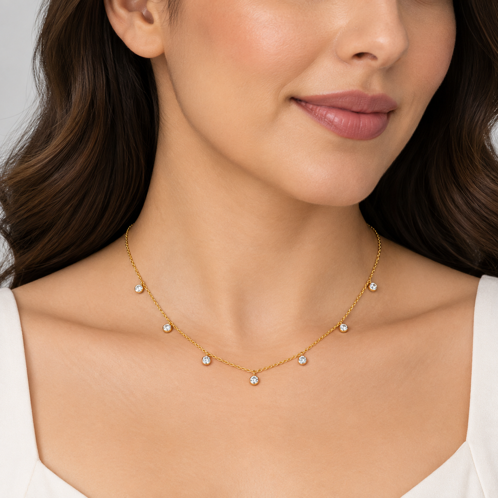 A close-up of a woman wearing a delicate gold necklace with five small clear gemstone drops spaced along the chain. The necklace is shown against her neck and upper chest, with her hair and white top visible.