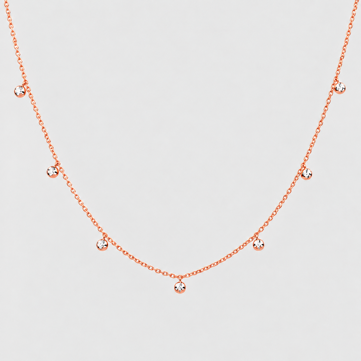 A delicate rose gold chain necklace featuring five small, clear round-cut gemstones spaced along the chain, displayed against a light gray background.