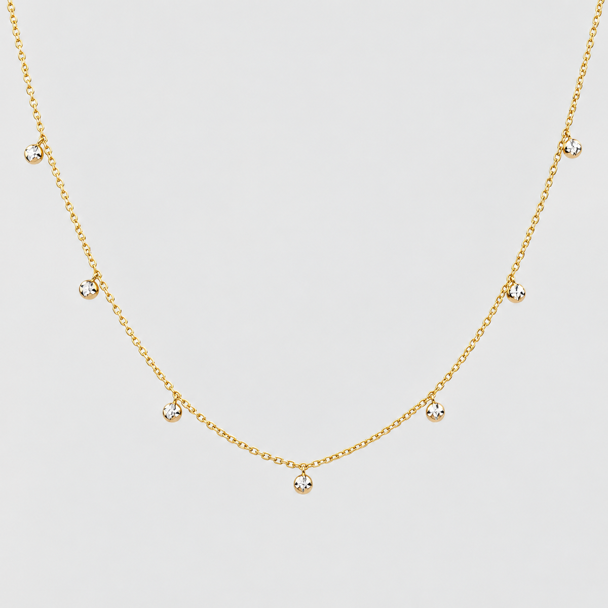 A delicate gold chain necklace featuring seven small, clear round-cut gemstones spaced evenly along the chain, displayed against a light gray background.