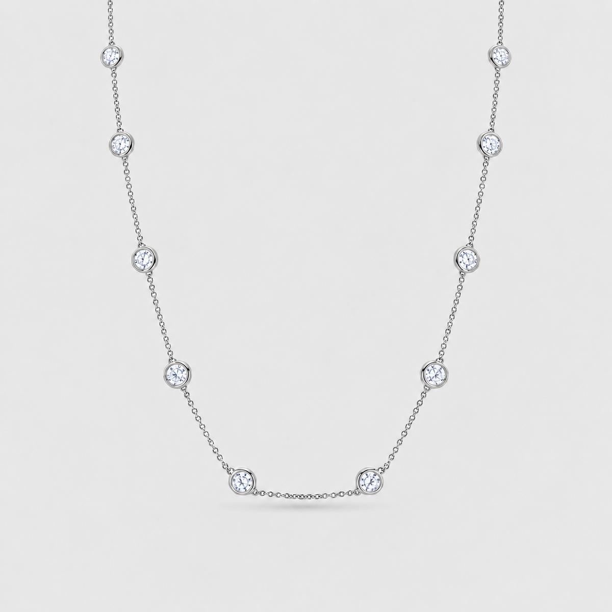 A delicate silver-toned chain necklace featuring seven evenly spaced round bezel-set clear gemstones, displayed against a light gray background.