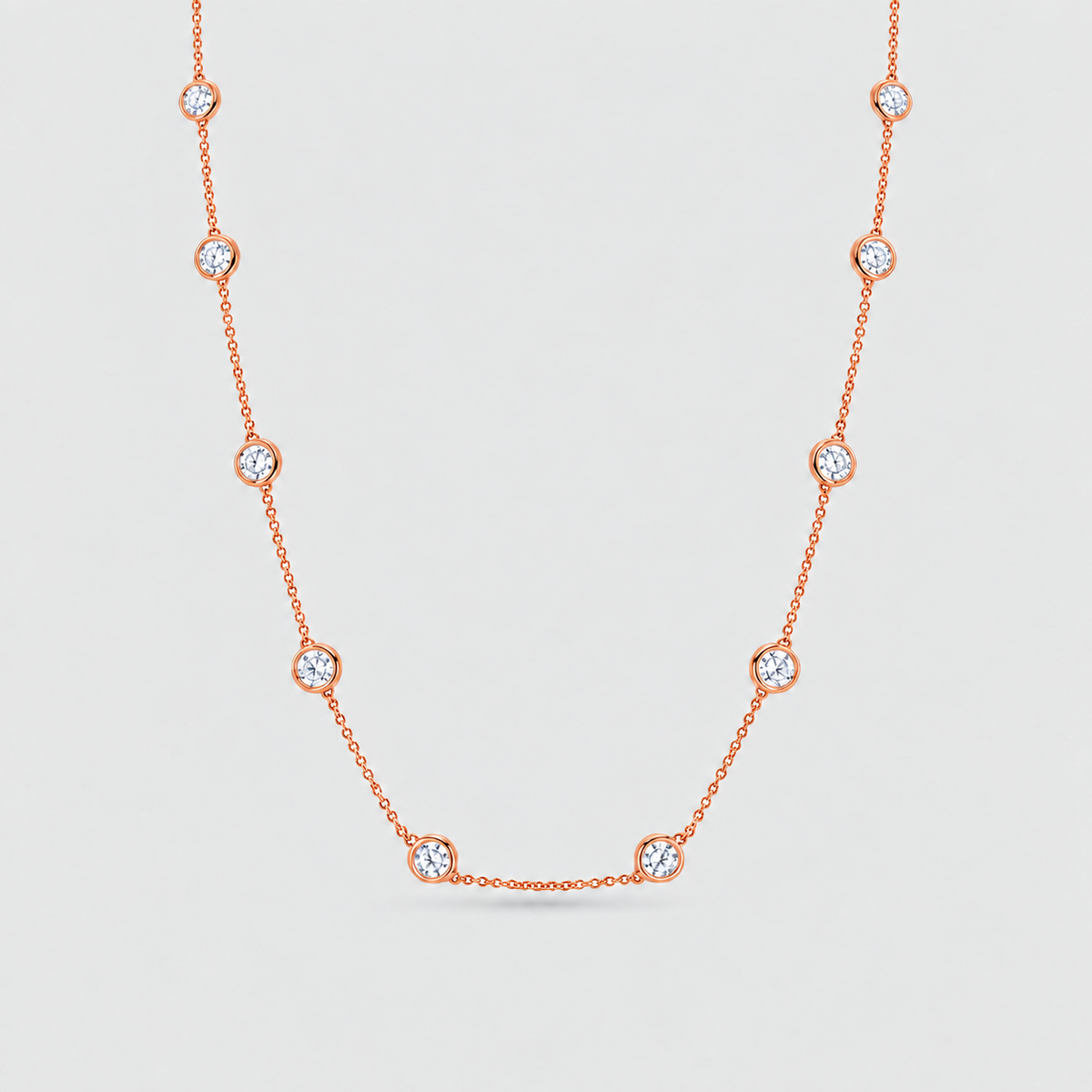 A delicate rose gold chain necklace featuring nine small, bezel-set round clear gemstones spaced evenly along the chain, displayed against a light gray background.