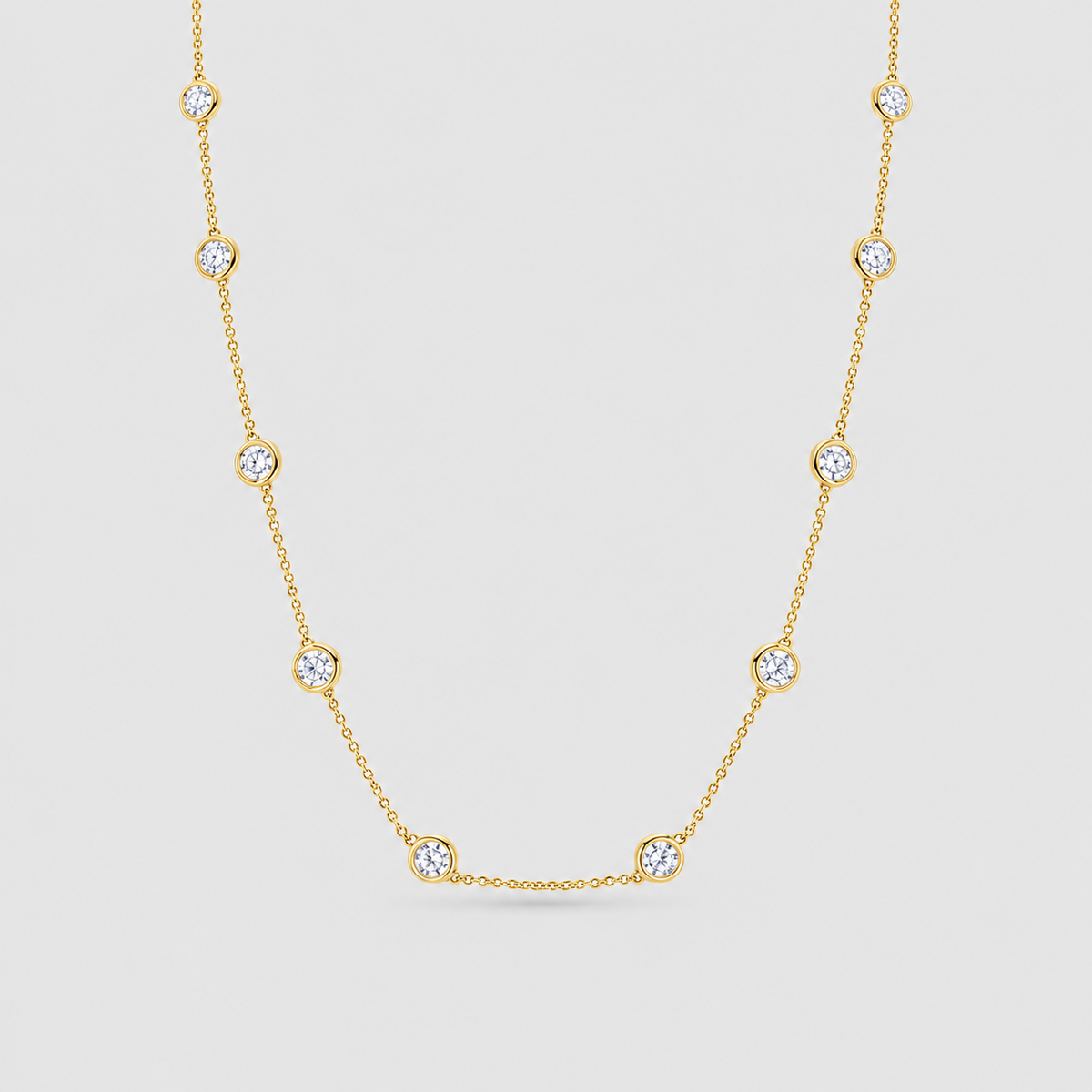 A delicate yellow gold chain necklace featuring nine small, bezel-set round clear gemstones spaced evenly along the chain, displayed against a light gray background.