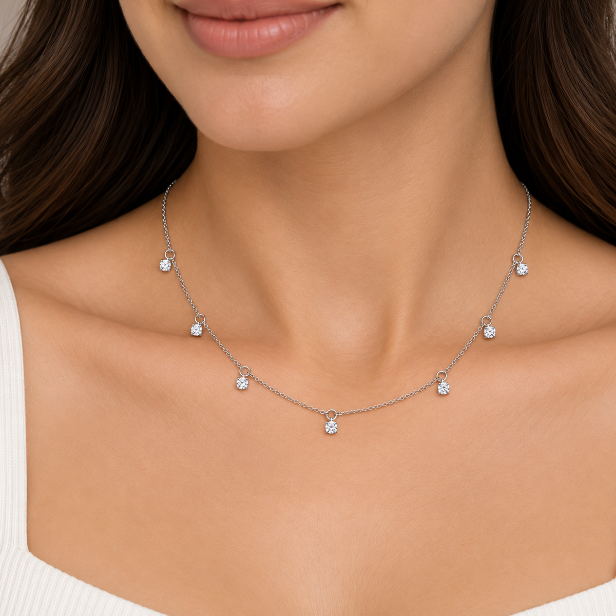 A close-up of a model's neck and upper chest wearing a delicate silver chain necklace with five small clear gemstone drops.