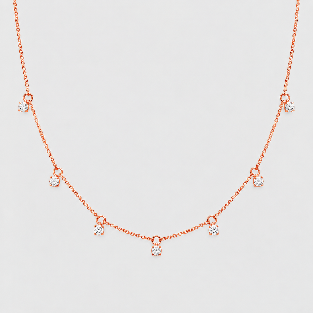 A delicate rose gold necklace featuring a fine chain with seven small, spaced bow-shaped charms, each adorned with a clear round-cut gemstone, set against a solid light gray background.