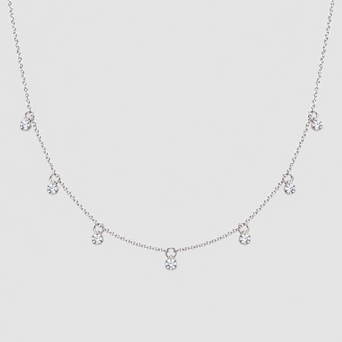 A delicate silver-tone chain necklace featuring five small clear crystal charms spaced along the front, displayed against a solid light gray background.