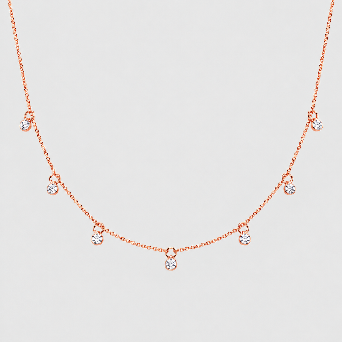 A delicate rose gold necklace featuring a fine chain with small, spaced-out teardrop-shaped crystal charms, displayed against a light gray background.