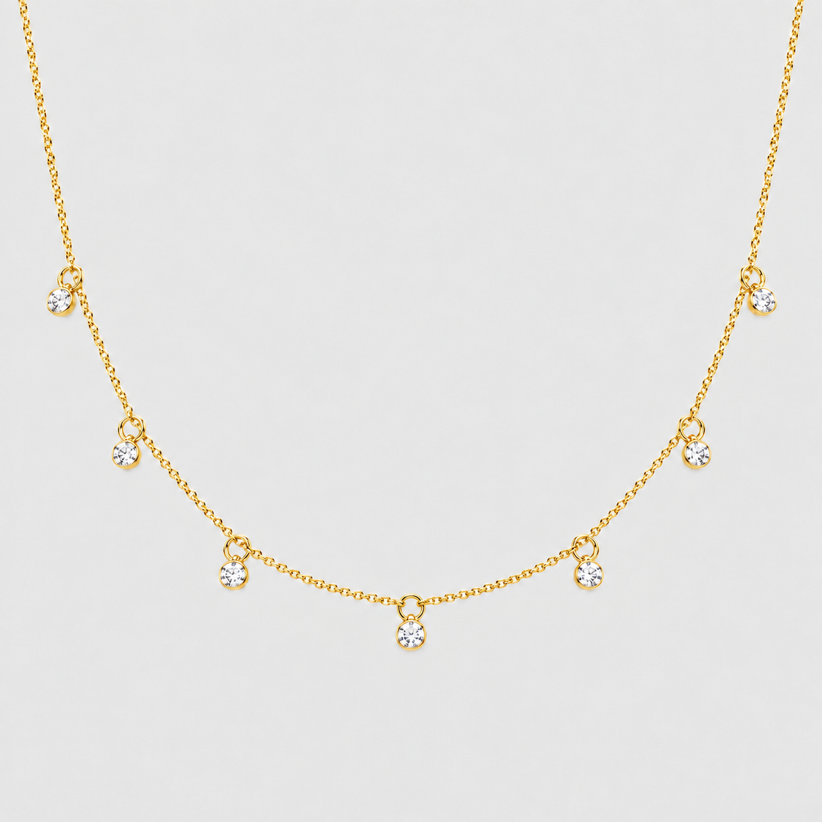 A delicate gold chain necklace featuring five small, clear round-cut gemstones spaced along the chain, displayed against a light gray background.