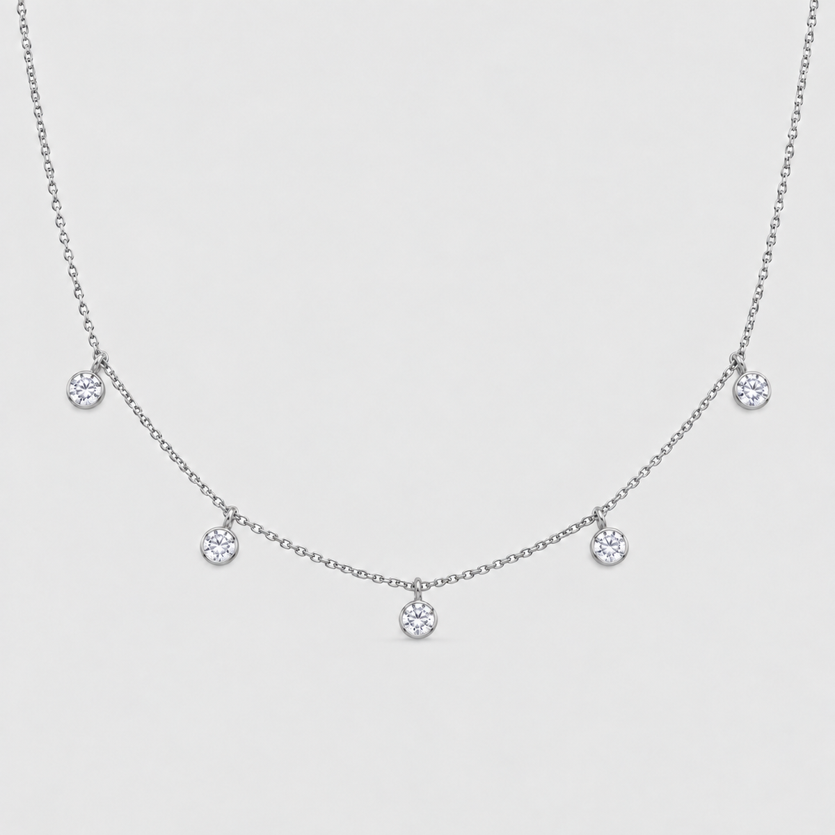 A delicate silver-toned chain necklace featuring five small, bezel-set round clear gemstones spaced along the chain, displayed against a light gray background.