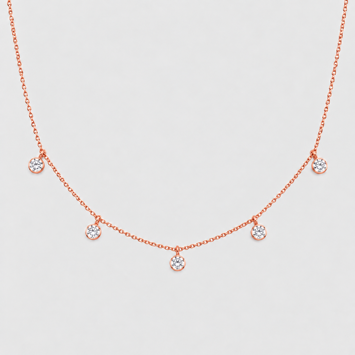 A delicate rose gold necklace featuring five small, round clear gemstones spaced along the chain, displayed against a light gray background.
