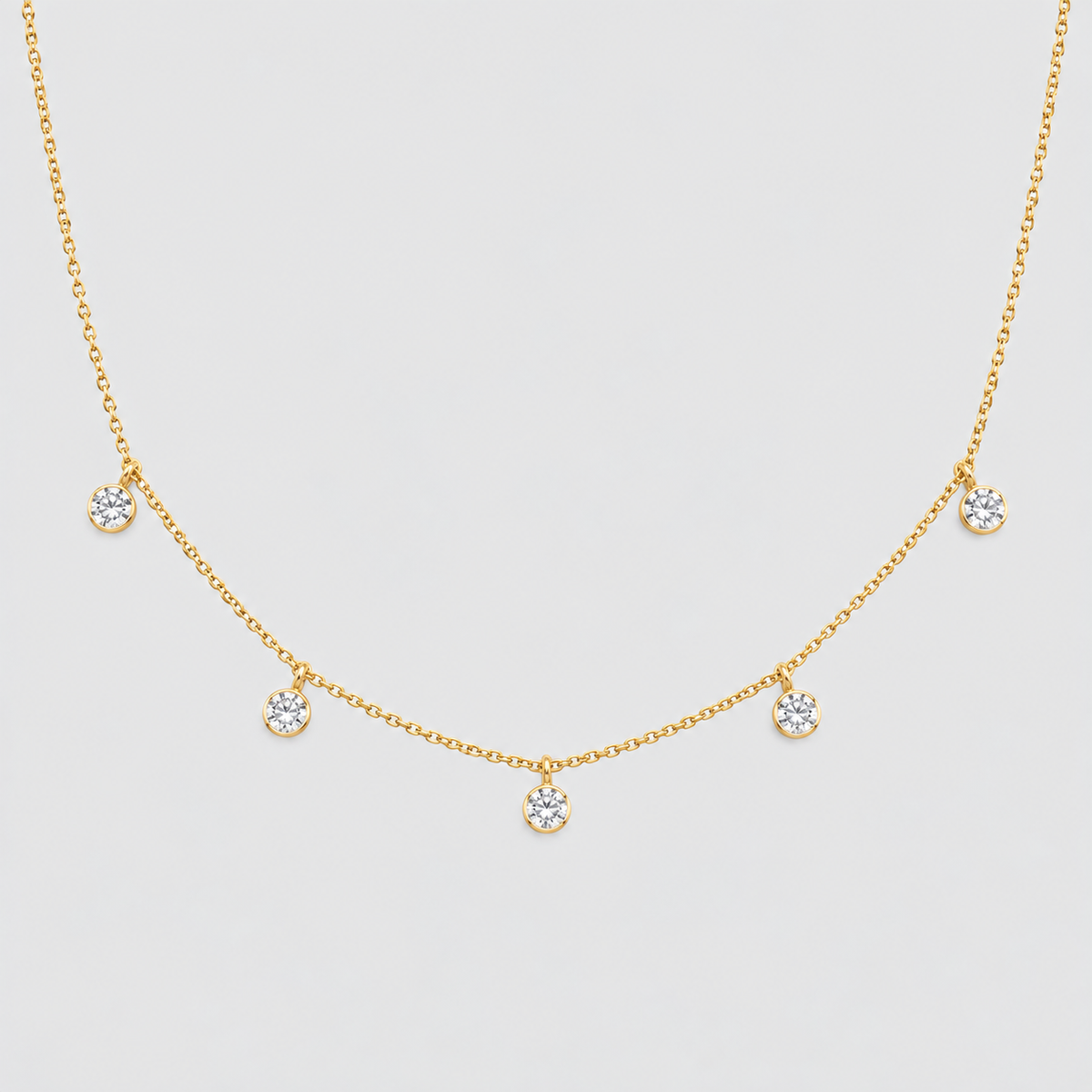 A delicate gold chain necklace featuring five small, round bezel-set clear gemstones spaced evenly along the chain, displayed against a light gray background.
