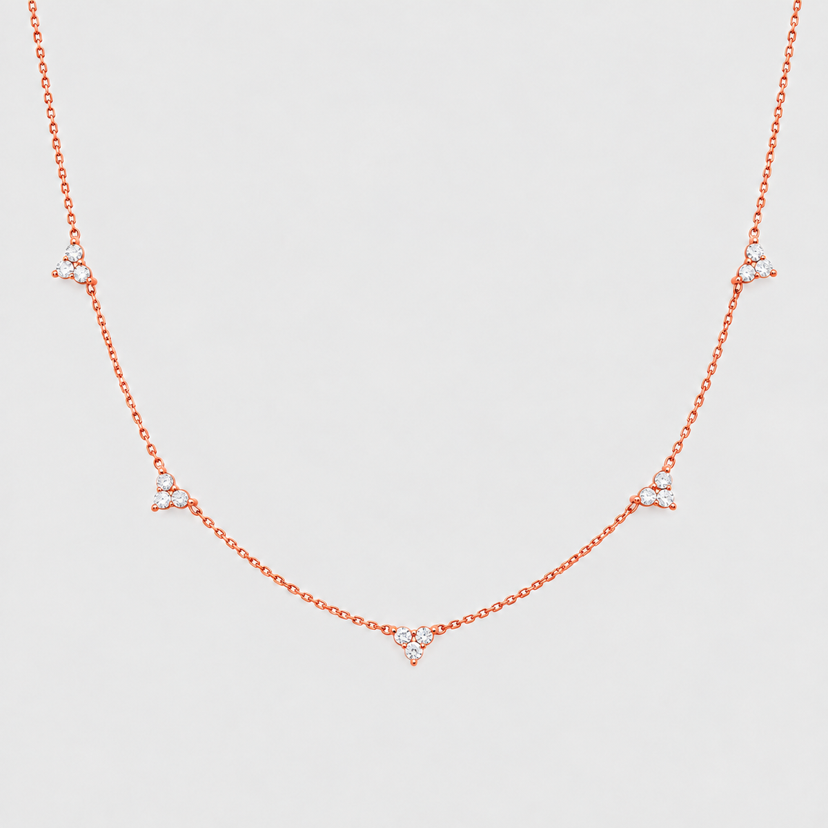 A delicate rose gold chain necklace featuring five small triangular clusters of clear stones spaced along the chain, displayed against a light gray background.
