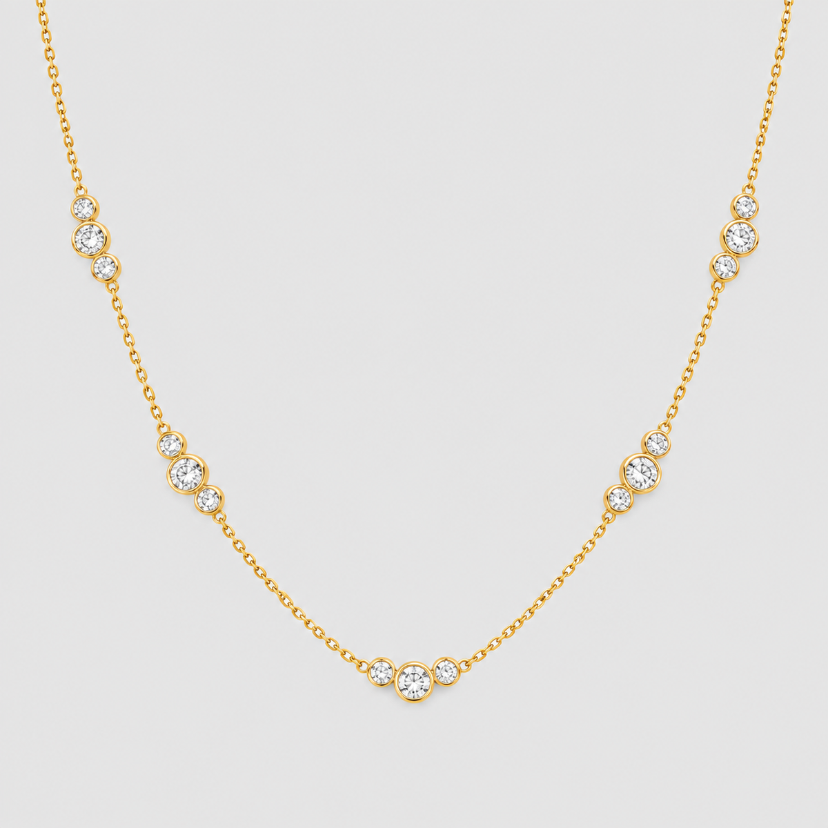 A delicate yellow gold chain necklace featuring multiple small bezel-set round clear stones spaced along the chain, displayed against a solid light gray background.