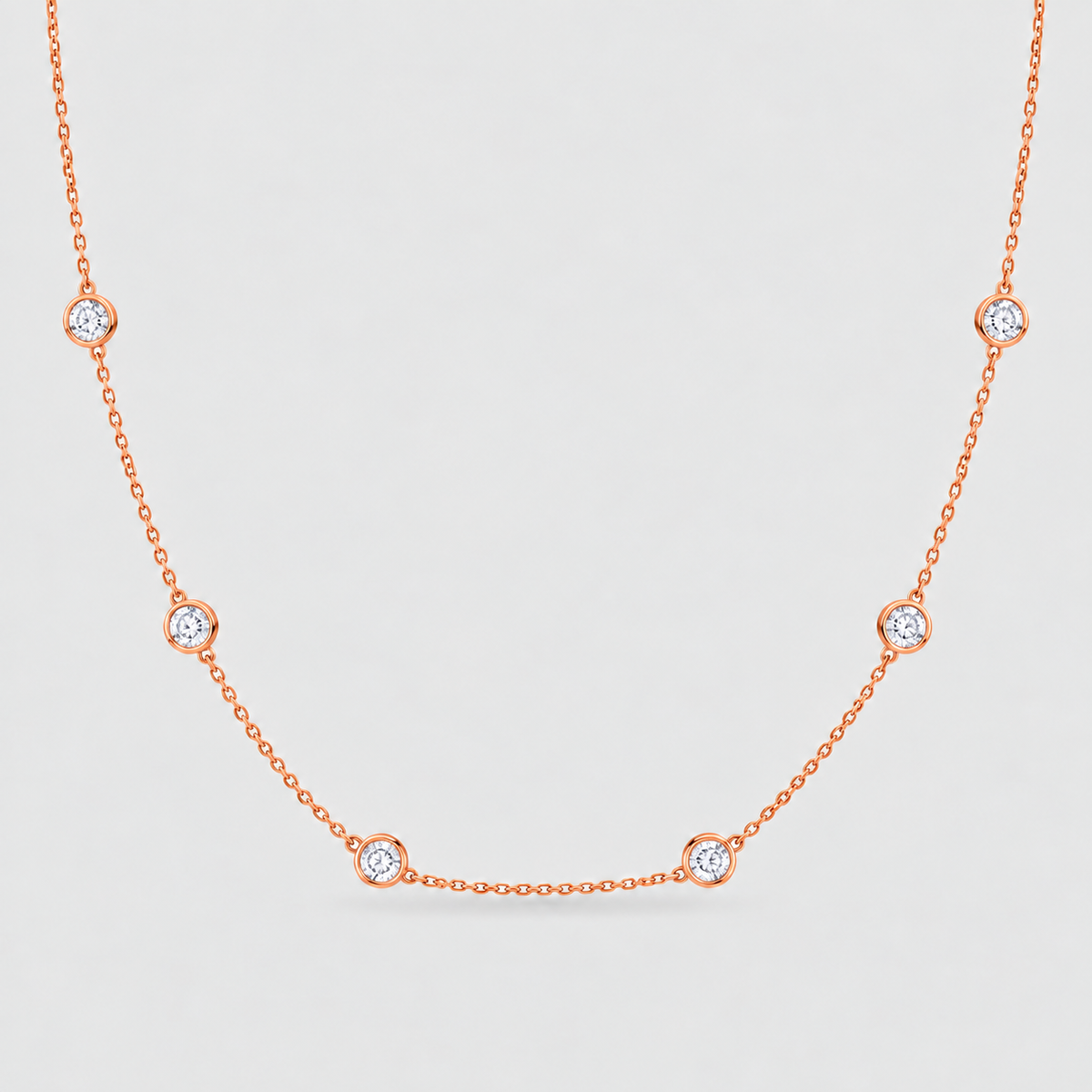 A delicate rose gold chain necklace featuring seven small, bezel-set round clear gemstones spaced evenly along the chain, displayed against a light gray background.