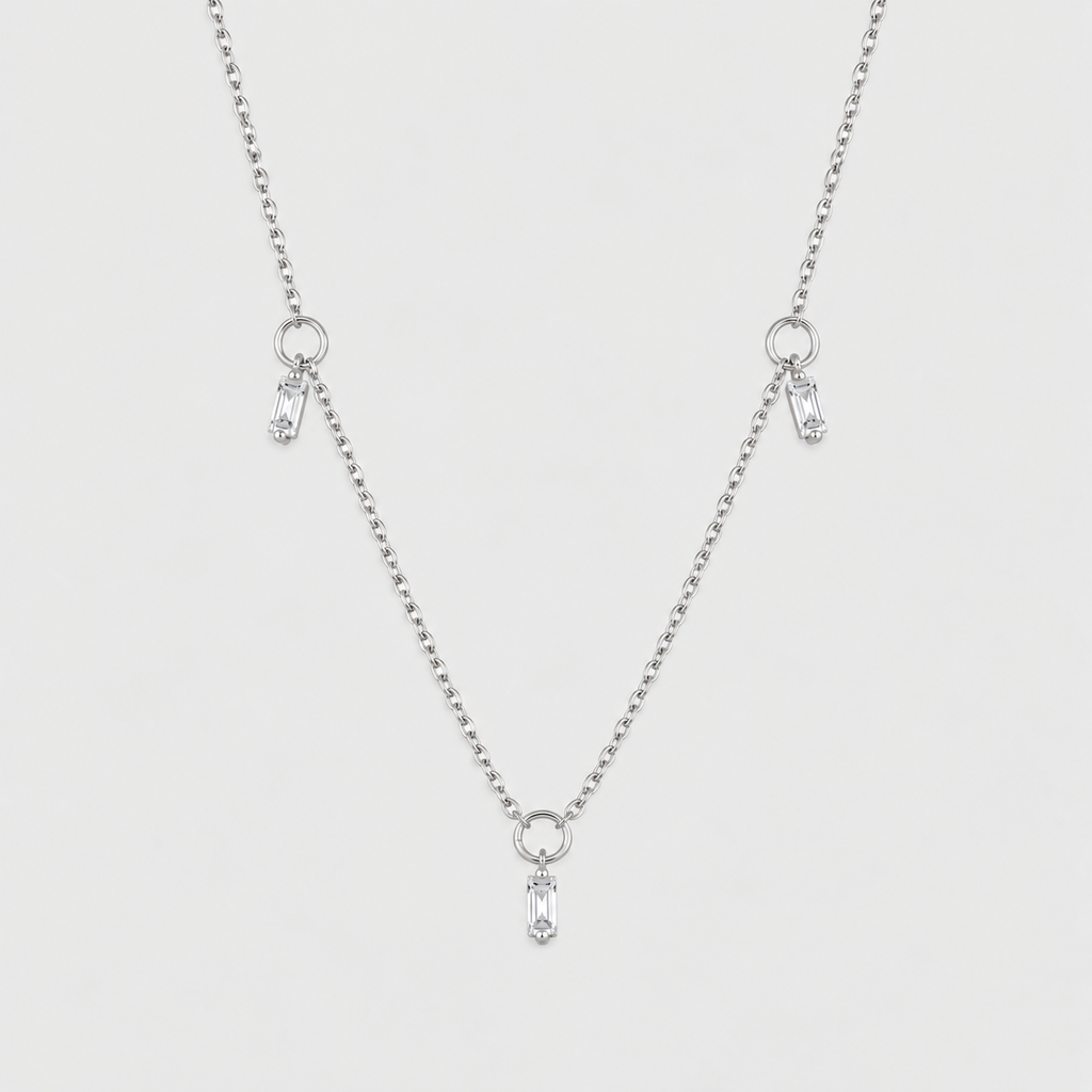 A delicate silver ball chain necklace featuring three small dangling charms, each with a clear teardrop-shaped crystal, set against a solid light gray background.