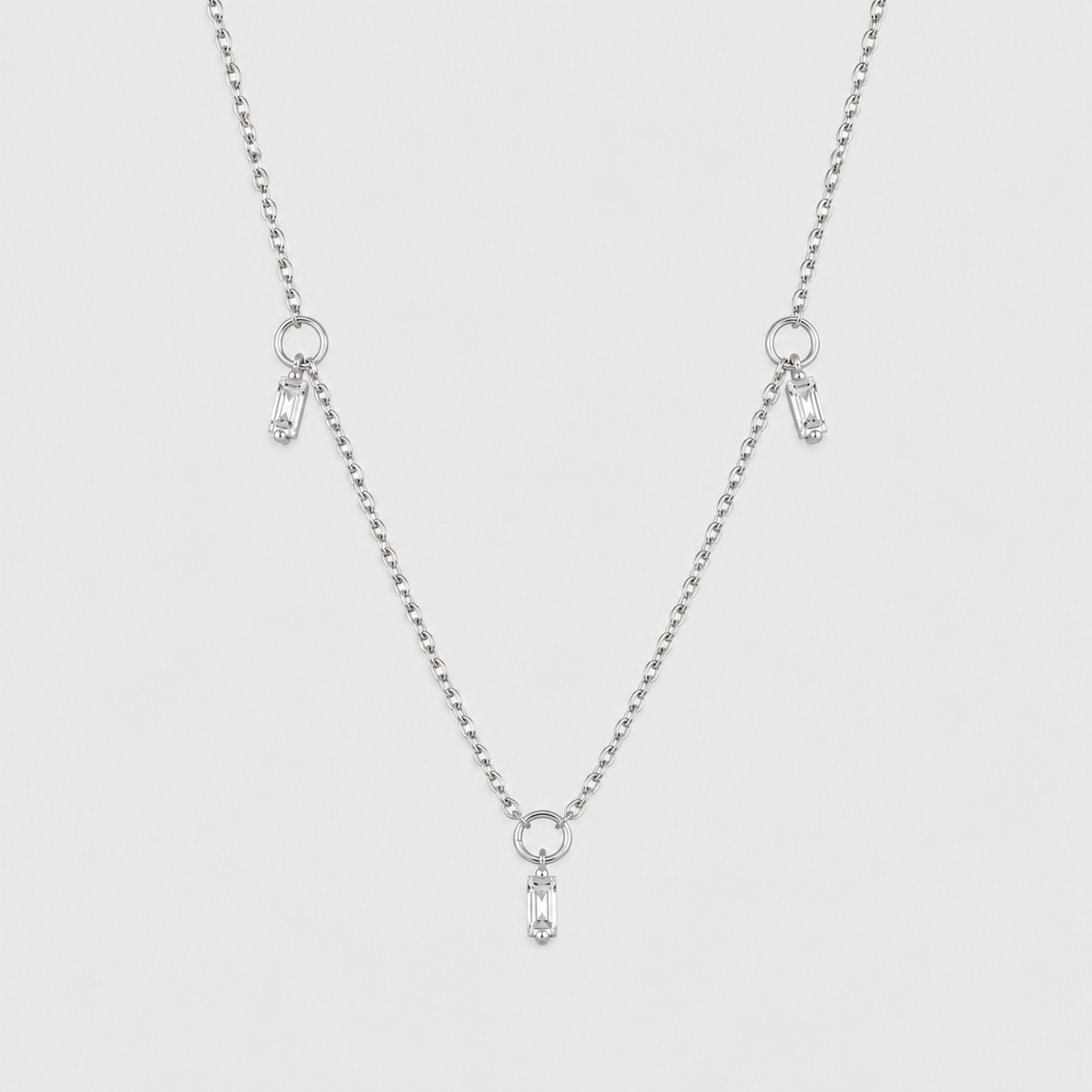 A delicate silver ball chain necklace featuring three small dangling charms, each with a clear teardrop-shaped crystal, set against a solid light gray background.
