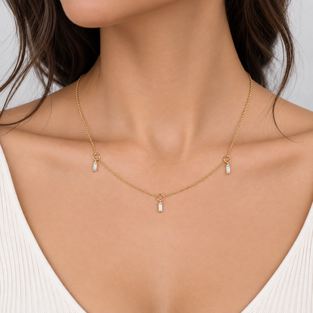 A close-up of a woman wearing a delicate gold chain necklace with three small white rectangular drop charms, styled with a white ribbed top.