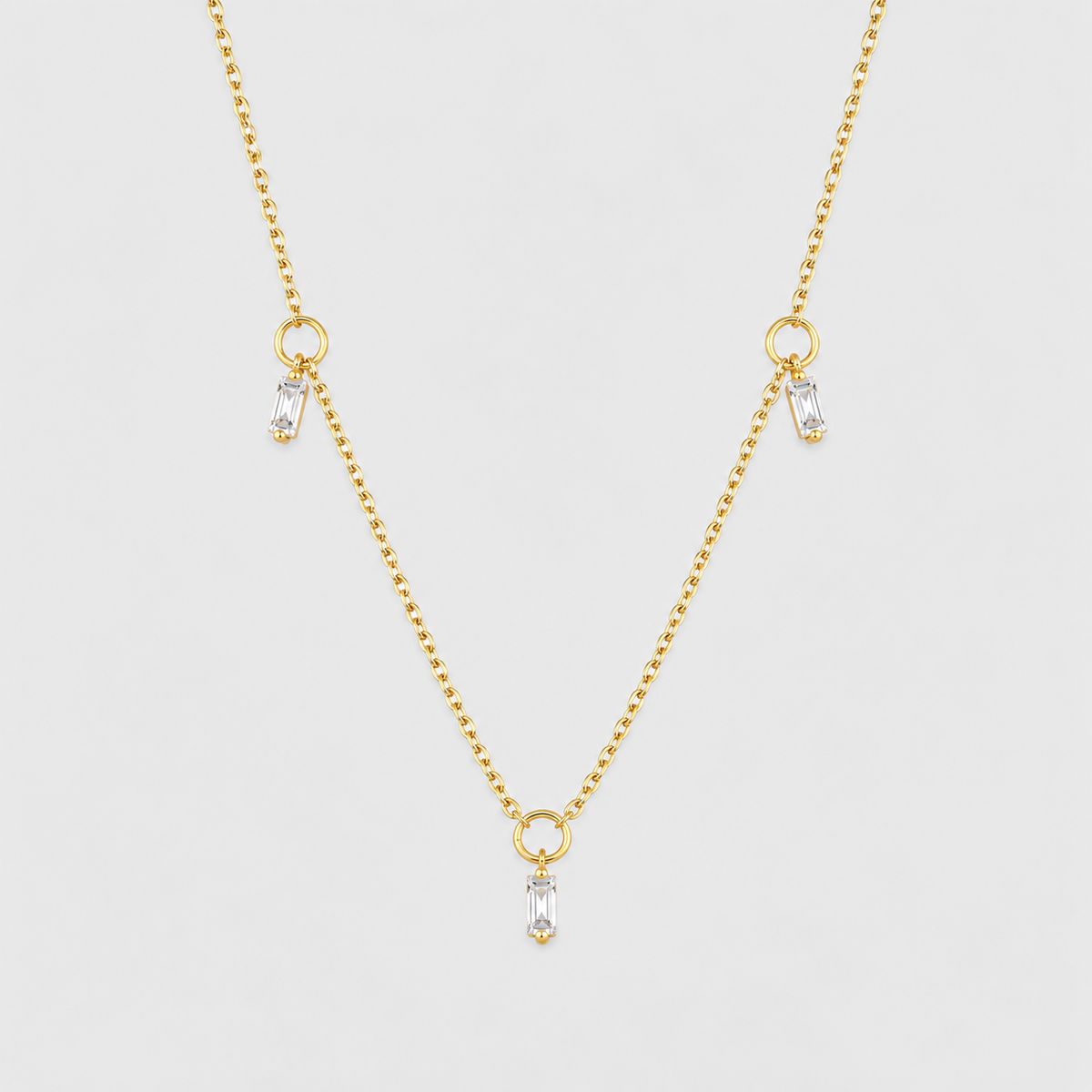 A delicate gold chain necklace featuring three small teardrop-shaped clear crystals hanging from gold rings at the center and two side locations, displayed against a light gray background.
