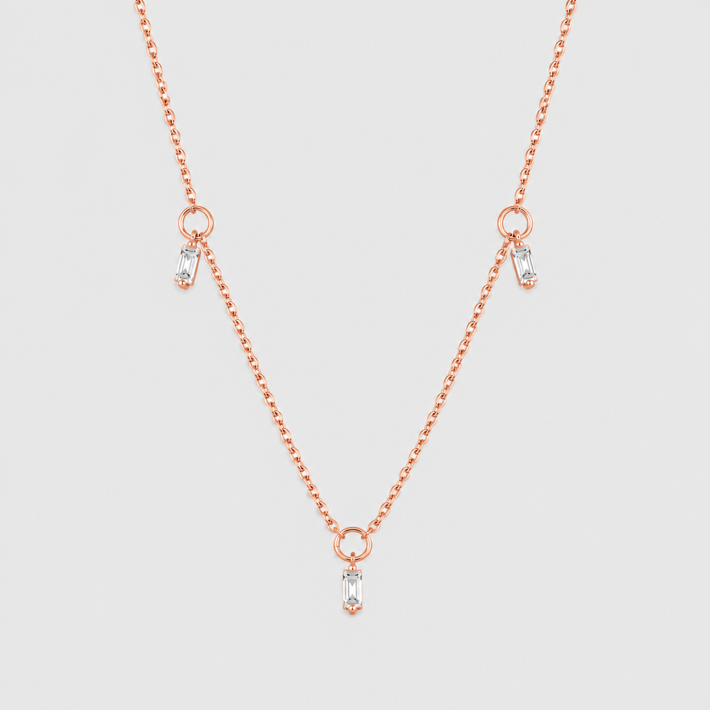 A delicate rose gold chain necklace featuring three small drop pendants, each adorned with a clear baguette-cut gemstone, set against a solid light gray background.