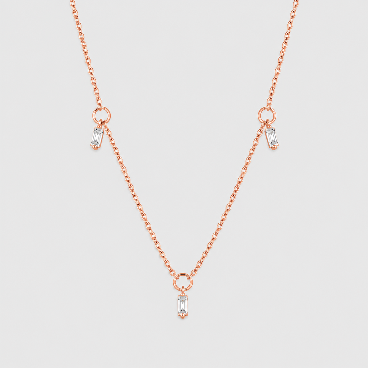 A delicate rose gold chain necklace featuring three small drop pendants, each adorned with a clear baguette-cut gemstone, set against a solid light gray background.