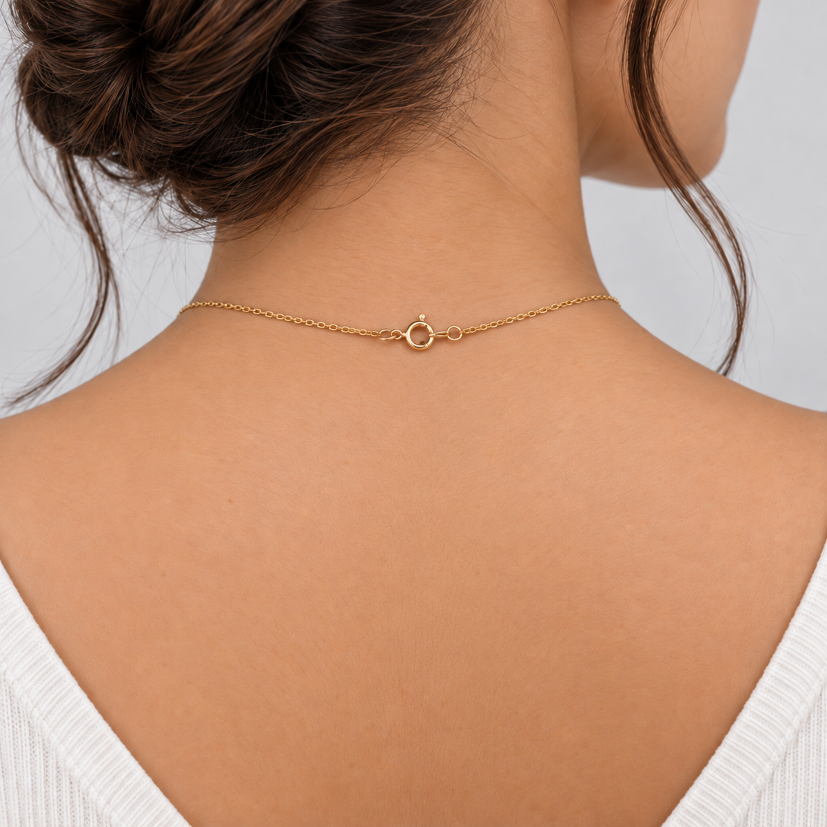 A close-up back view of a woman wearing a delicate gold chain necklace with a small circular ring clasp, shown against a light gray background.
