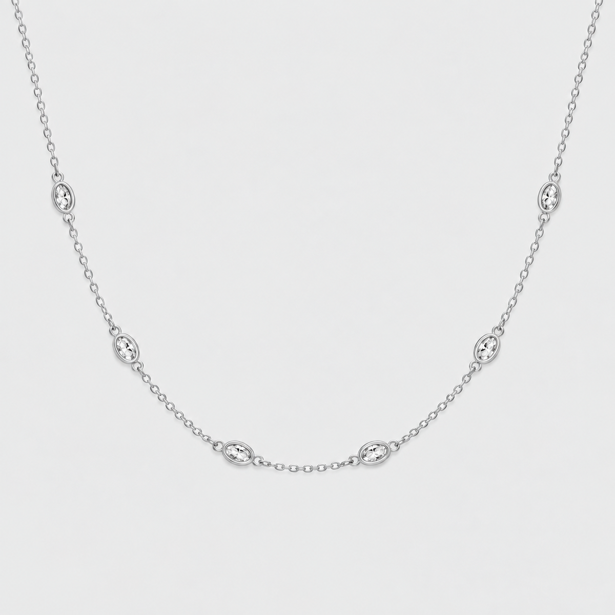 A delicate silver chain necklace featuring five small oval bezel-set clear stones, displayed against a clean, light gray background.