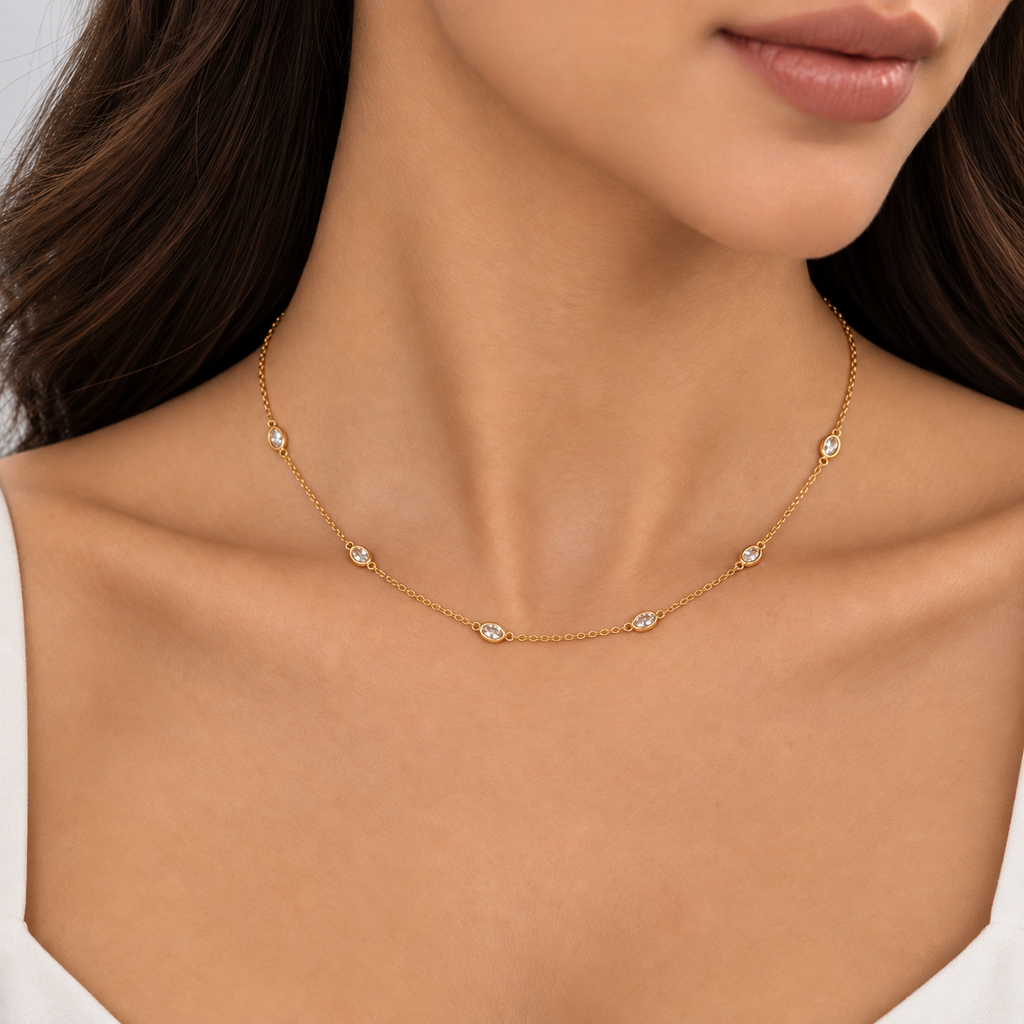 A close-up of a model wearing a delicate gold chain necklace with small oval clear gemstone accents, shown against her neck and upper chest.