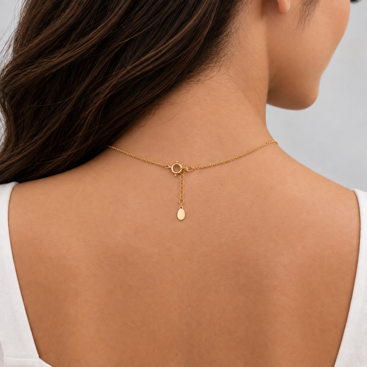 A close-up back view of a woman wearing a delicate gold chain necklace with a small teardrop pendant and extension chain, shown against her skin.