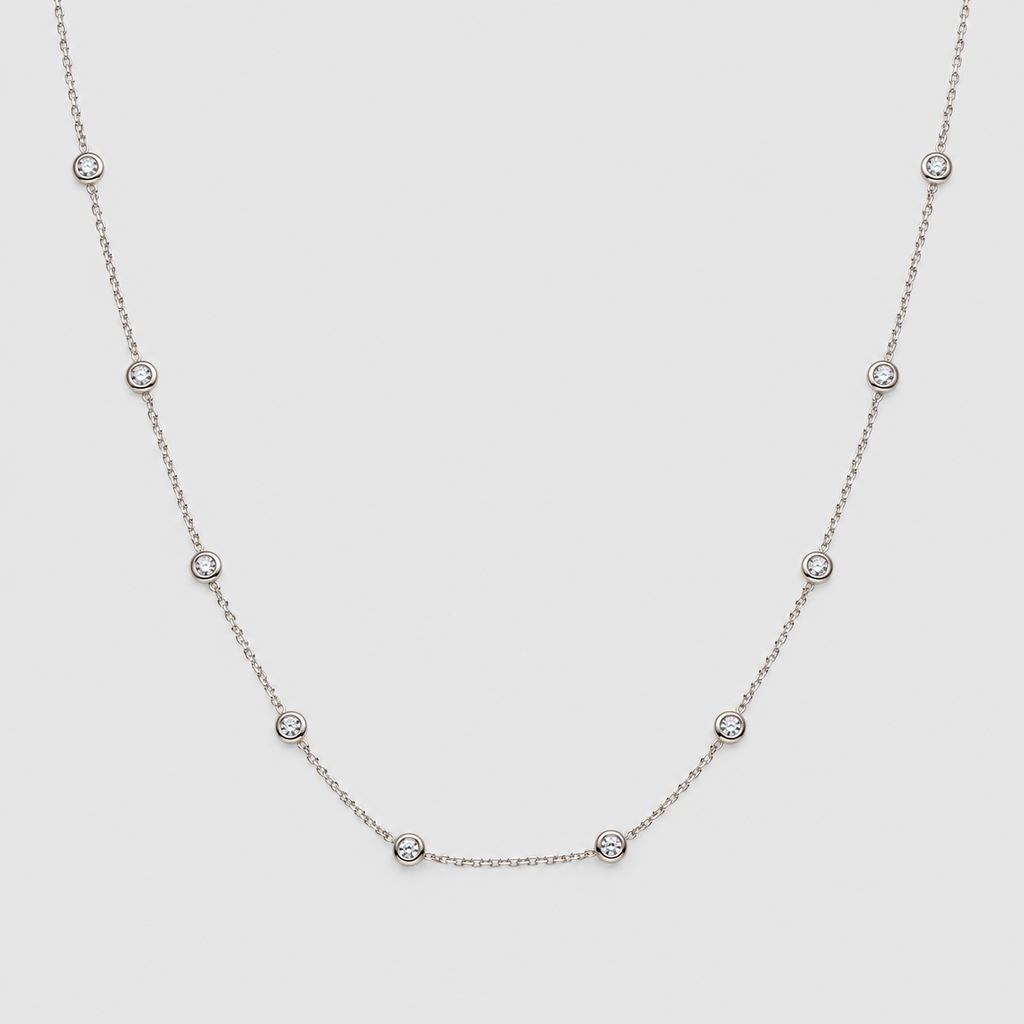 A delicate silver chain necklace featuring seven small, evenly spaced round bezel-set clear gemstones, displayed against a solid light gray background.