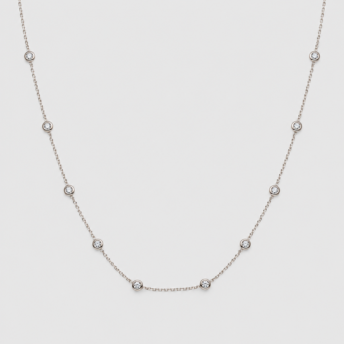 A delicate silver chain necklace featuring seven small, evenly spaced round bezel-set clear gemstones, displayed against a solid light gray background.