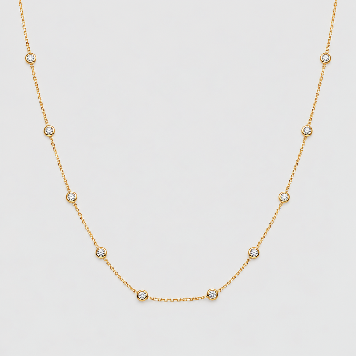 A delicate gold chain necklace featuring seven small, bezel-set round clear gemstones spaced evenly along the chain, displayed against a light gray background.