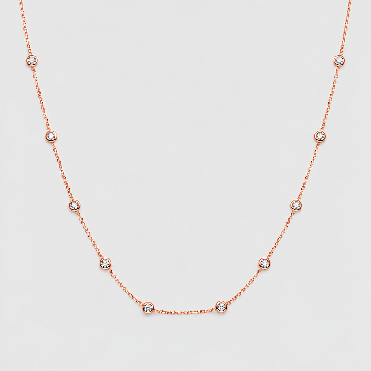 A delicate rose gold necklace featuring a fine chain and small, evenly spaced bezel-set round clear gemstones, displayed on a light gray background.