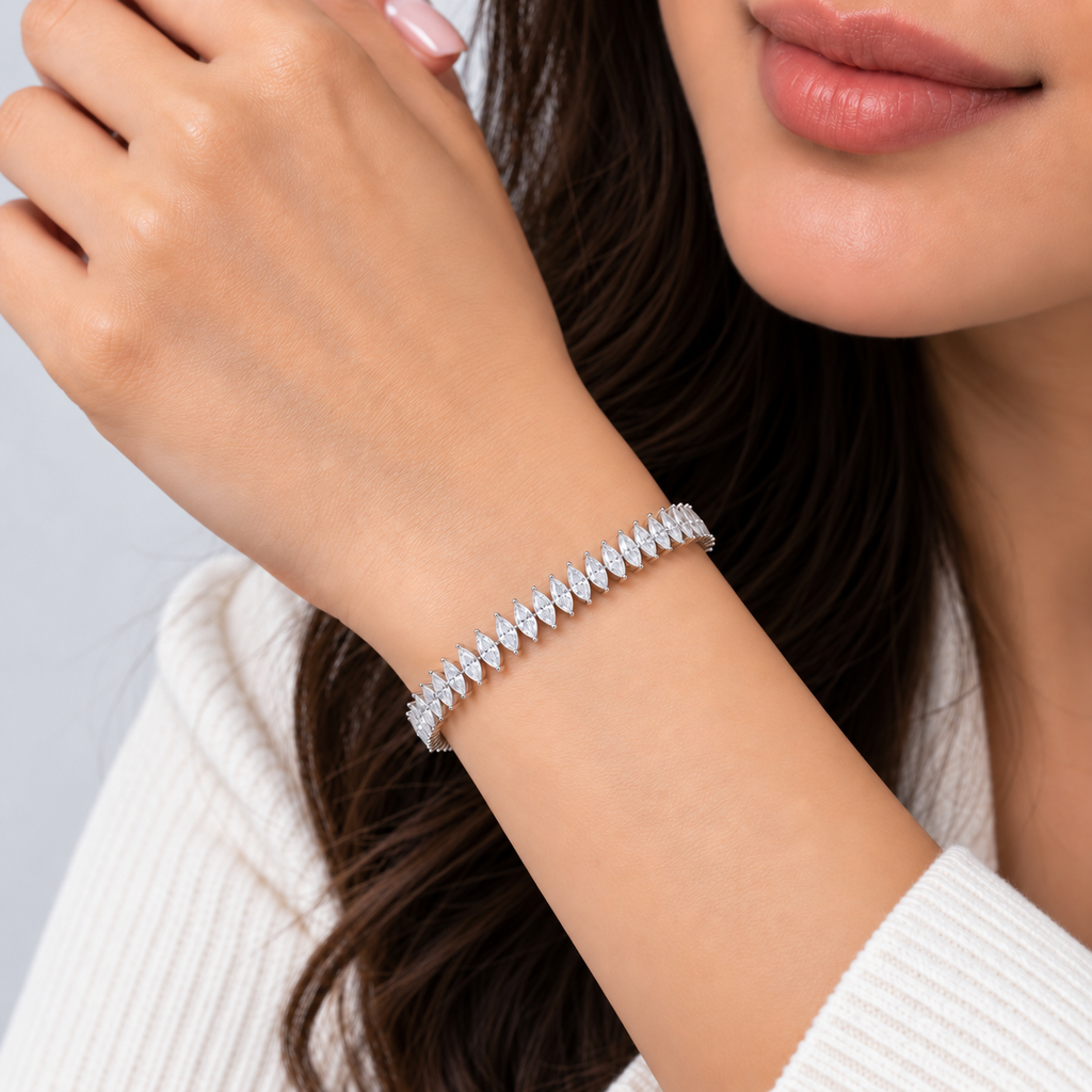 A close-up lifestyle shot of a woman wearing a sparkling silver tennis bracelet with marquise-cut stones. The image features her hand resting near her face, showcasing the bracelet on her wrist against a soft, light gray background.