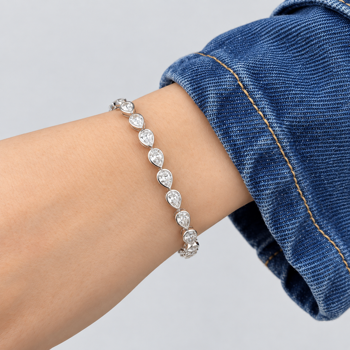 A silver-toned tennis bracelet with teardrop-shaped clear stones, shown worn on a person's wrist with a blue denim cuff visible.