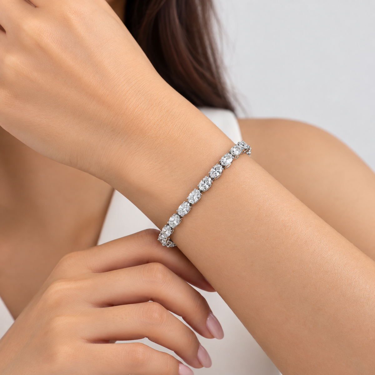 A close-up of a woman's wrist wearing a sparkling diamond tennis bracelet in a silver-tone setting, with her hands and arms visible against a clean light gray background.