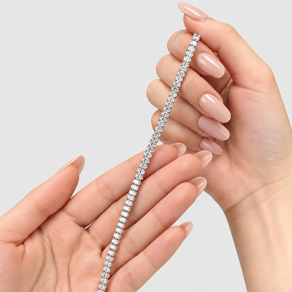 A close-up of a person's hands holding a delicate silver tennis bracelet with sparkling clear stones against a light gray background.