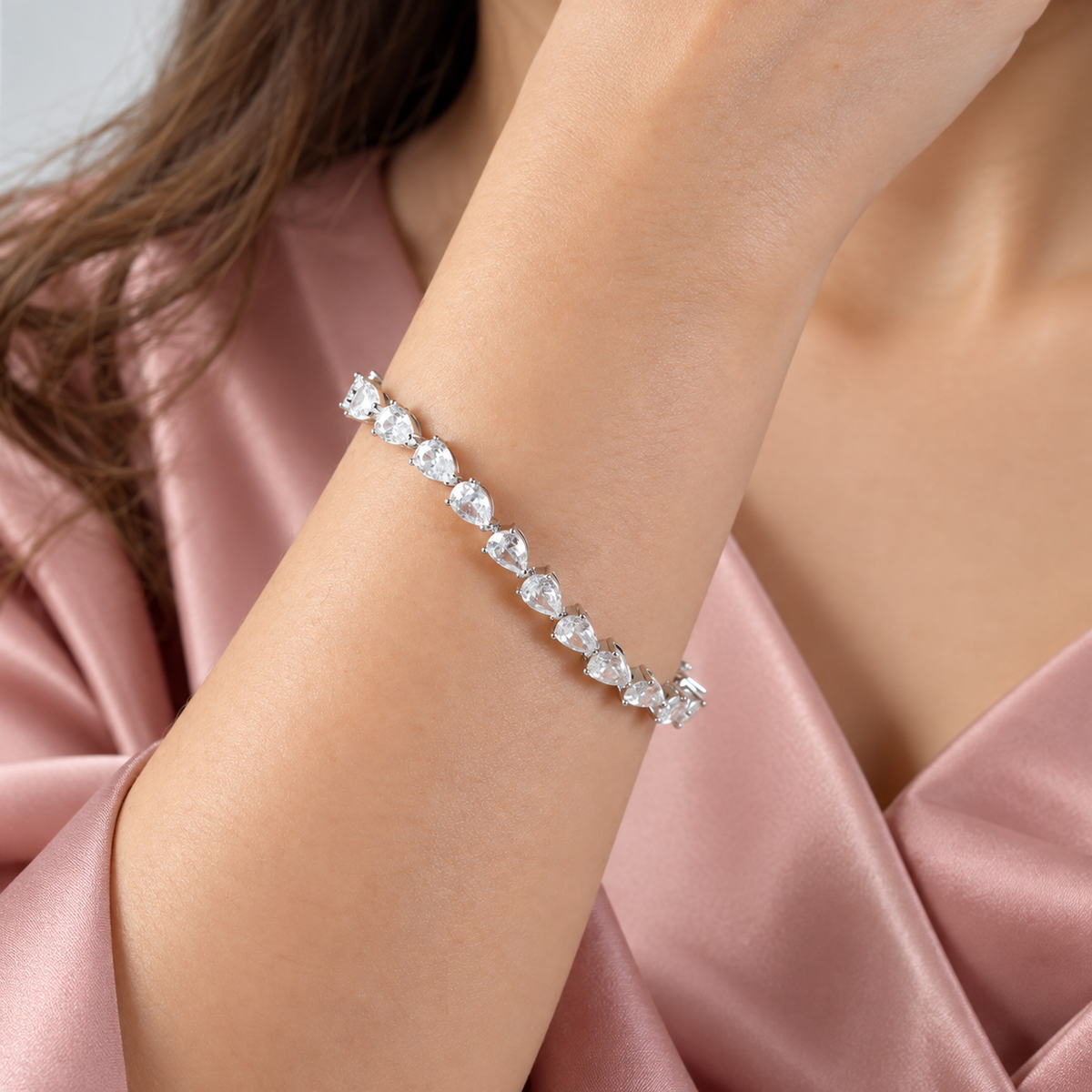 A close-up of a woman's wrist wearing a silver-toned tennis bracelet with clear teardrop-cut gemstones, styled with a soft pink satin robe.