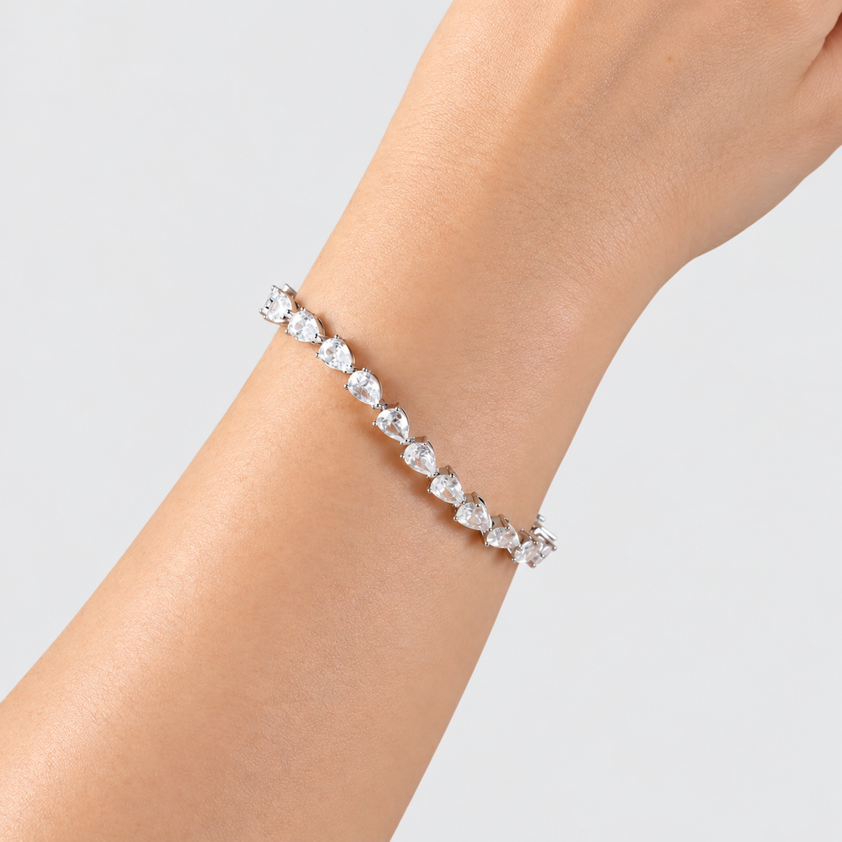 A silver-toned tennis bracelet with clear heart-shaped gemstones worn on a person's wrist against a light gray background.