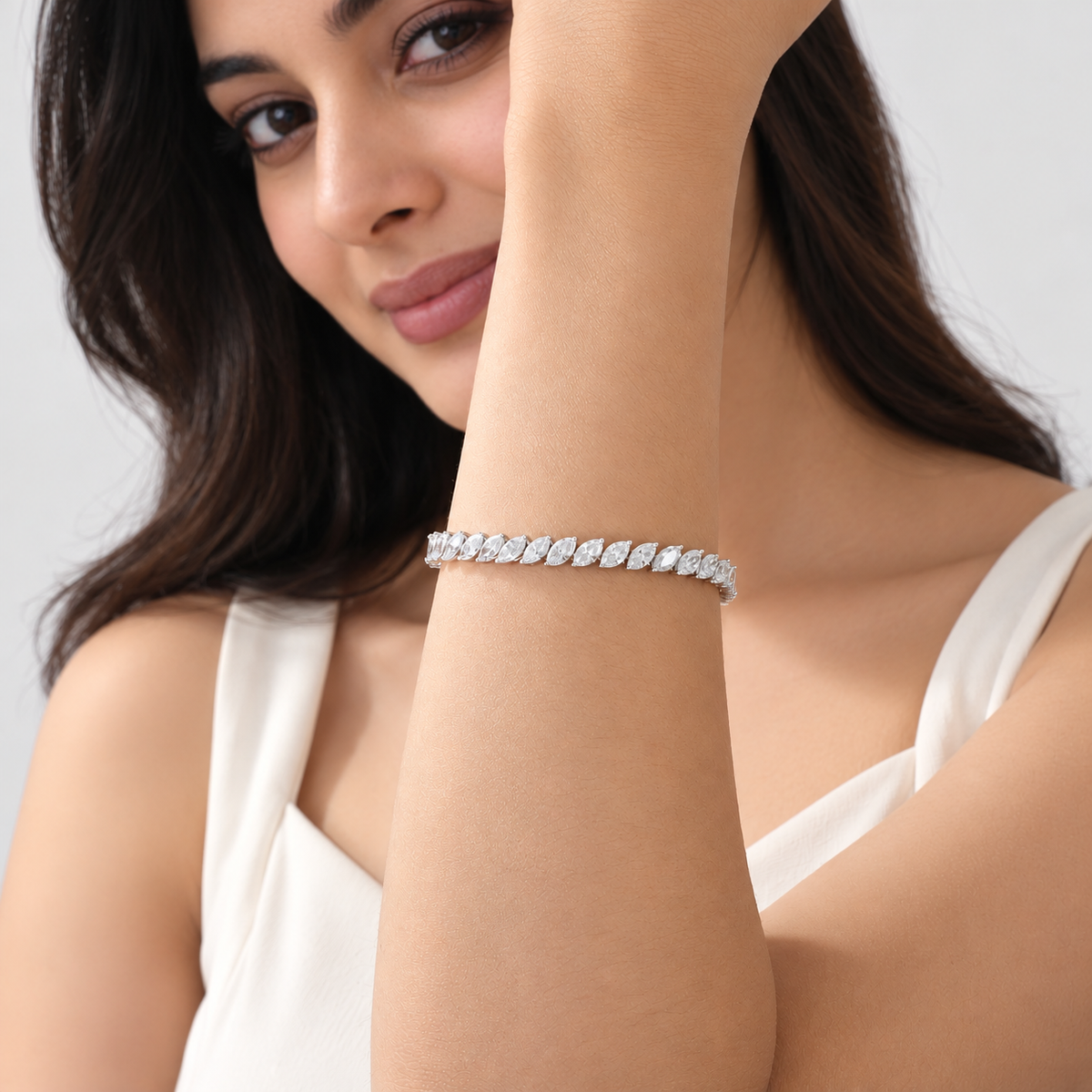 A woman wearing a silver tennis bracelet with marquise-cut stones, shown in a close-up portrait against a light gray background.